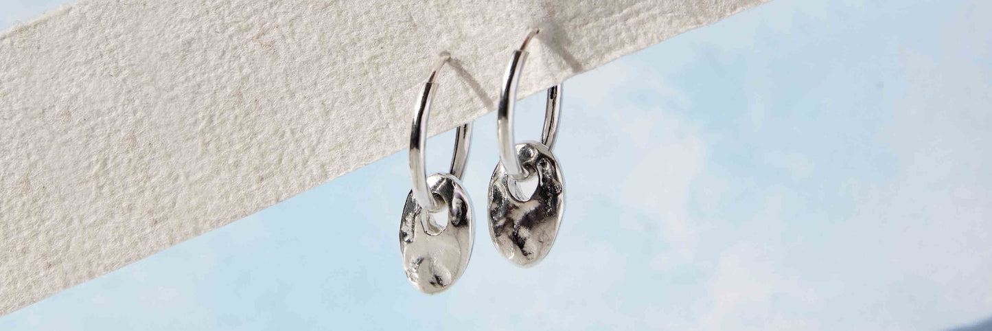 Image of two silver huggie hoop earrings with hammered oval silver charms susopended froma  strip of textured white paper