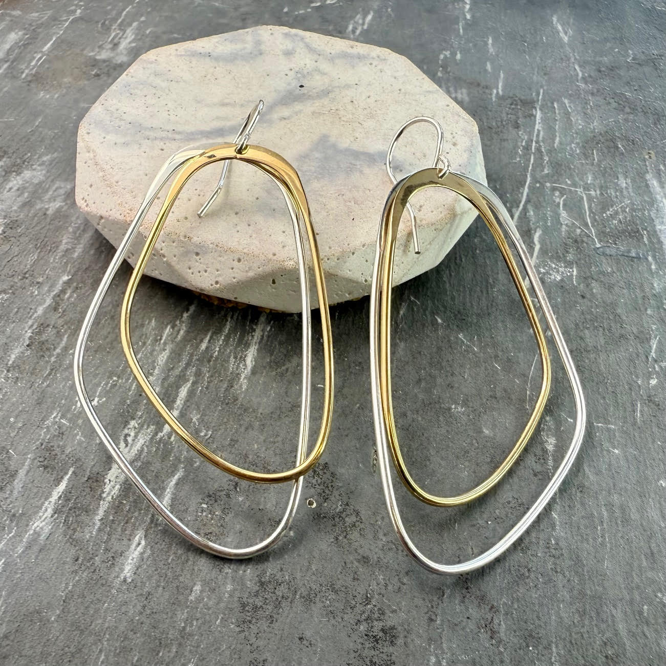A pair of silver and gold double hoop earrings displayed on a textured surface with a natural stone.