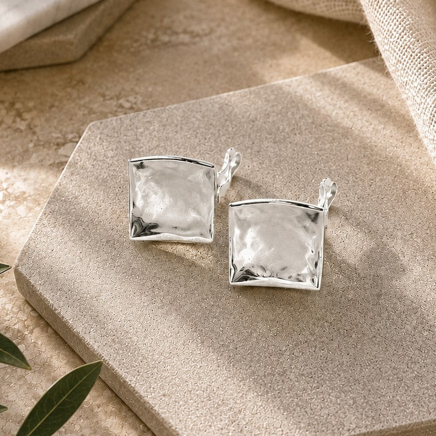 A pair of silver-toned square clip-on earrings with clear faceted stones, displayed on a textured beige stone slab with linen and marble props in a warm neutral setting.