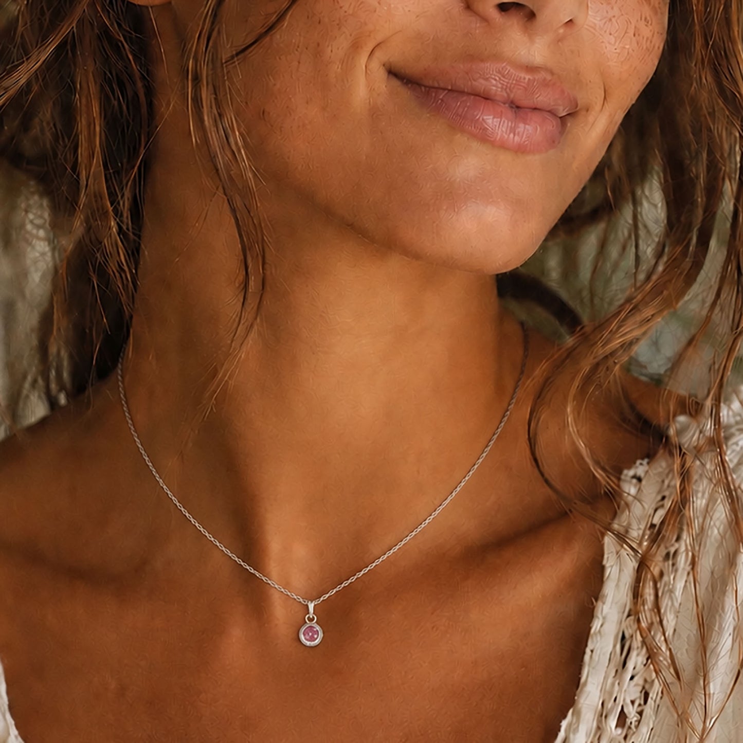 A close-up lifestyle shot of a woman wearing a delicate silver chain necklace with a small round pendant featuring a pink gemstone.